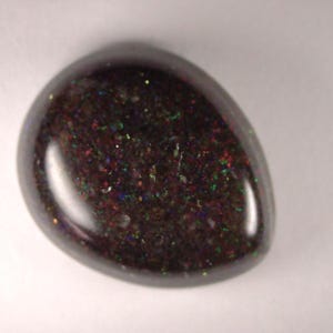 May include: A polished, teardrop-shaped black opal gemstone. The opal has a dark base color with iridescent flecks of red, green, and blue, creating a shimmering effect. The stone is smooth and glossy, reflecting light.