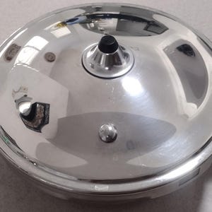 May include: A shiny, stainless steel pressure cooker lid with a black handle and a black knob. The lid has a reflective surface and two black handles with the text "DUROMATIC" on them.