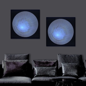 May include: Two framed abstract art prints with a blue and white circular design on a black background. The prints are hanging on a grey wall above a black sofa with cushions.