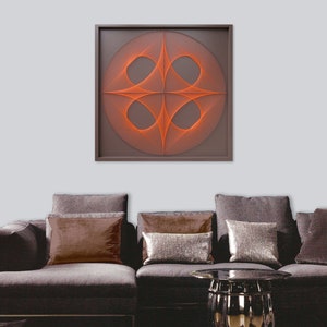 May include: A framed wall art piece with a geometric design made of orange string. The design is a circle with four intersecting lines that create a pattern of four ovals. The art is hanging on a wall above a gray sofa with brown pillows.