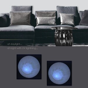 May include: Two square framed wall art pieces with a circular design. The design is made of white thread on a purple background. The art is shown hanging above a grey sofa with cushions. The same art is shown again in a darker setting with the thread glowing blue under UV light.