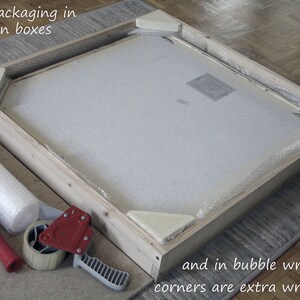 May include: A wooden box with a white foam insert and bubble wrap. The box is being taped shut with a red tape dispenser. The text "safe packaging in wooden boxes" and "and in bubble wrapper corners are extra wrapped" is visible in the image.