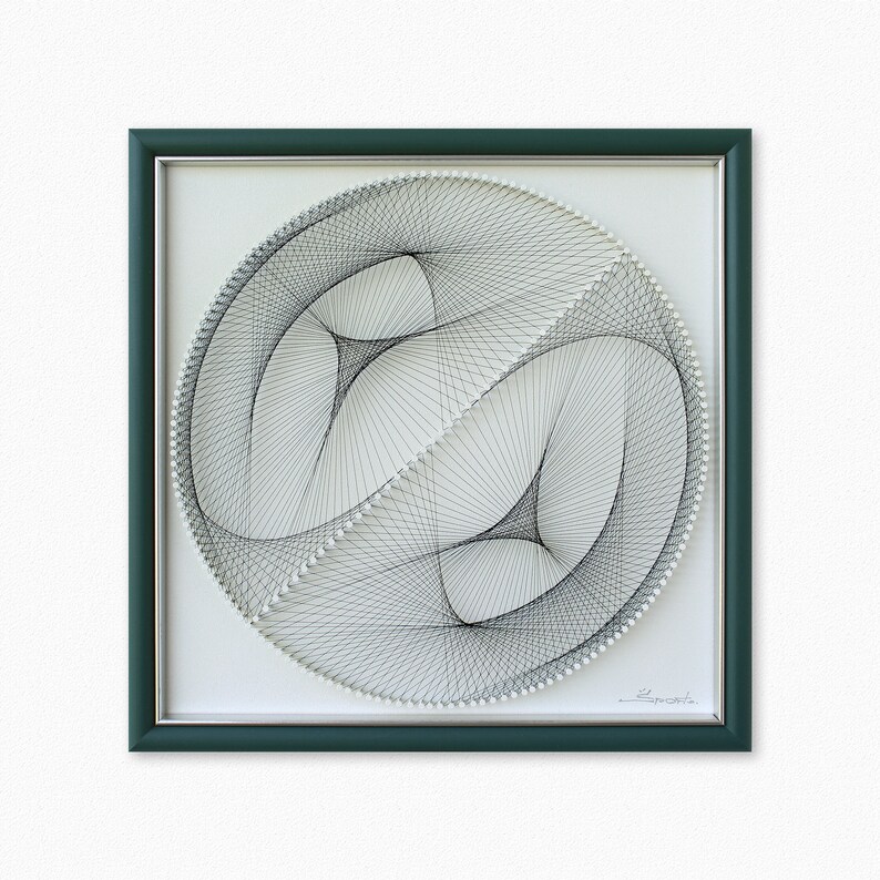 Puede incluir: Una pieza de arte de hilo enmarcada con un dise&ntilde;o en blanco y negro de dos c&iacute;rculos superpuestos. El dise&ntilde;o se crea con hilo envuelto alrededor de clavos en un patr&oacute;n circular. El marco es de color verde oscuro.