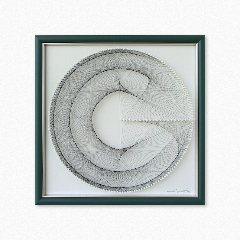 Puede incluir: Una obra de arte enmarcada que presenta un dise&ntilde;o de arte de hilo negro y blanco de un c&iacute;rculo con una l&iacute;nea curva en su interior. El marco es de color verde oscuro.