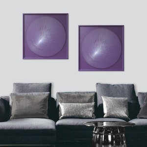 May include: Two square framed wall art pieces with a purple background and a white circular design in the centre. The art is hanging on a white wall above a grey sofa with silver cushions.
