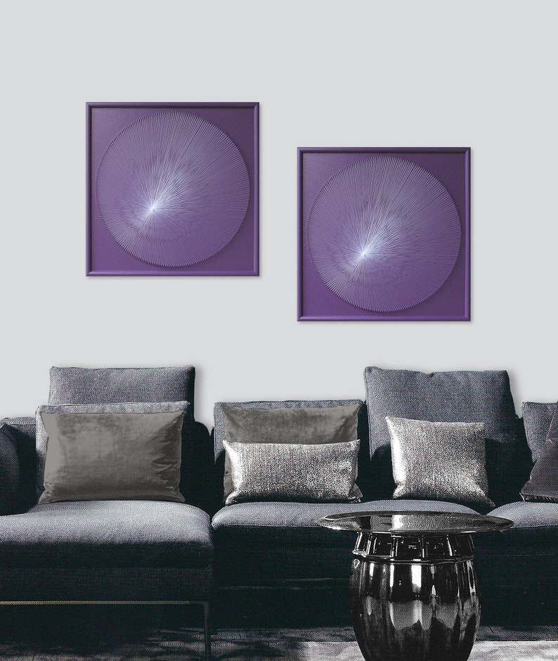 May include: Two square framed wall art pieces with a purple background and a white circular design in the centre. The art is hanging on a white wall above a grey sofa with silver cushions.