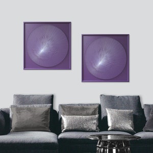 May include: Two square wall art pieces with purple frames and white thread designs in the shape of a circle. The art pieces are hanging on a white wall above a grey sofa with grey throw cushions.