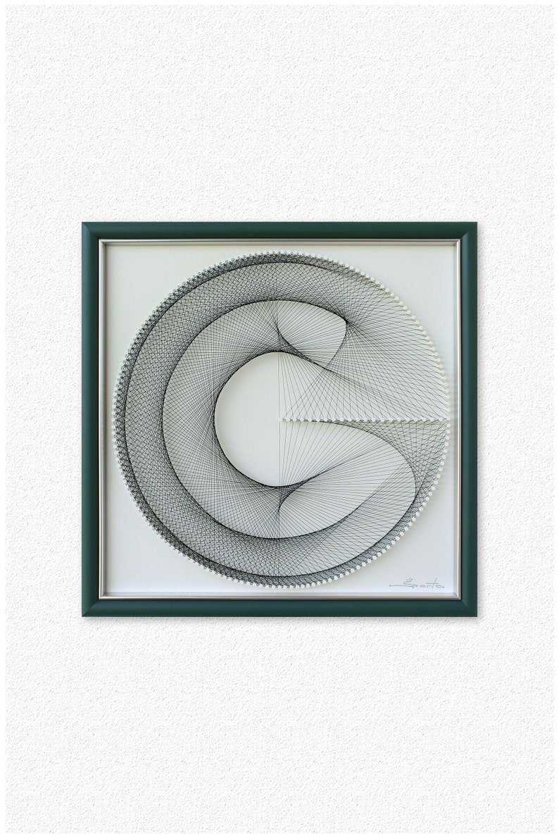 Puede incluir: Una pieza de arte de hilo en blanco y negro enmarcada en un marco verde oscuro. El arte de hilo representa un c&iacute;rculo con una l&iacute;nea curva en su interior, creando un dise&ntilde;o geom&eacute;trico. La pieza est&aacute; firmada "L. Saputo" en la esquina inferior derecha.
