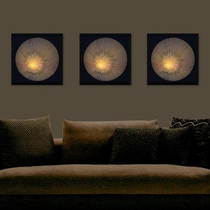 May include: Three black framed wall art pieces with gold and white abstract designs. The art pieces are hanging on a brown wall above a brown sofa with cushions.