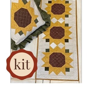 Può includere: Un kit per la trapunta con un motivo a girasole. Il kit comprende pezzi di tessuto in tonalità di giallo, marrone e verde, insieme a un tessuto di fondo bianco. Il kit è etichettato "kit".