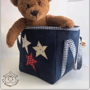 Può includere: Una borsa portaoggetti in tessuto blu con applicazioni a stella bianche e rosse e manici in nastro a quadri. Un orsacchiotto marrone è seduto all'interno della borsa.