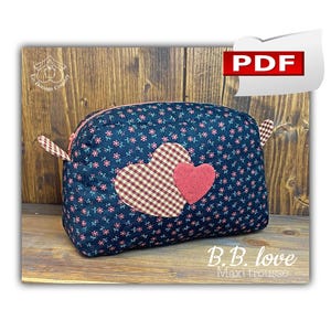 May include: A dark blue fabric makeup bag with a zipper closure. The bag has two hearts on the front, one red and one plaid. The text "B.B. love Maxi trousse" is printed below the hearts.