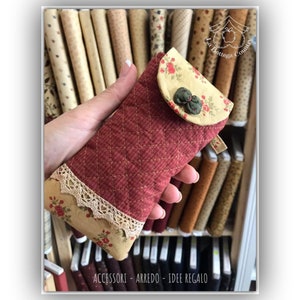 Könnte beinhalten: Eine gesteppte Handyhülle mit Blumenmuster und einem Klappverschluss. Die Hülle wird in einer Hand vor einem Hintergrund aus Stoffrollen gehalten.