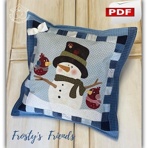 Può includere: Cuscino decorativo quadrato con un pupazzo di neve con cappello a cilindro nero, sciarpa e due uccelli rossi. Il cuscino ha un bordo patchwork blu e il testo "Frosty's Friends". Un fiocco bianco è sull'angolo superiore.
