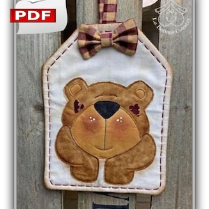 Può includere: Un appendiabiti a forma di orso in tessuto fatto a mano con un fiocco. L'orso è marrone con un naso nero e dettagli a forma di cuore. Lo sfondo è bianco con un bordo cucito e un fiocco a quadri. L'appendiabiti è attaccato a una superficie di legno.