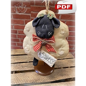 Può includere: Un ornamento a forma di pecora in feltro bianco con un viso nero, un fiocco a scacchi rosso e bianco e un supporto in legno marrone. L'ornamento ha un'etichetta che dice "Simply".