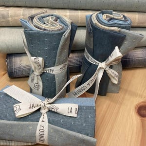 Può includere: Rotoli di tessuto nei toni del blu, grigio e beige, legati con nastri color crema. I nastri sono stampati con testo e piccole illustrazioni. I rotoli sono impilati su una superficie di legno, con più tessuto visibile sullo sfondo.