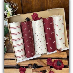 Può includere: Un set di cinque rotoli di tessuto in una scatola di cartone. Il tessuto è bianco con strisce rosse, rosso con stampa floreale bianca, bianco con pois rossi, rosso con stampa floreale bianca e bianco con bacche rosse. I rotoli di tessuto sono su una superficie di legno con bastoncini di cannella e fiori secchi.
