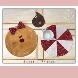 May include: Two round fabric coasters with a Christmas theme. One coaster is brown with a gingerbread man face and a red bow. The other coaster is white and red with a candy cane design. The text "Sweet Friends" is printed below the coasters.
