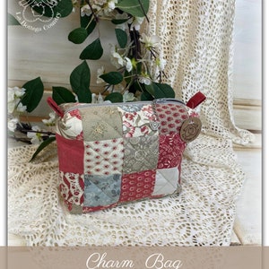 Può includere: Una borsa portaciondolo in patchwork rosso, bianco e blu con chiusura a zip. La borsa è appoggiata su una superficie di uncinetto bianca. La borsa ha un piccolo ciondolo attaccato al tiretto della zip. Il ciondolo è un cuore con un piccolo disegno di mela. La borsa è etichettata "Charm Bag".