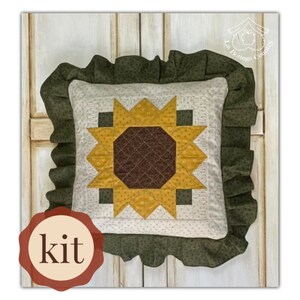 Può includere: Un kit per un cuscino a forma di girasole in tessuto verde e bianco. Il cuscino ha un bordo arricciato e il girasole è realizzato in tessuto giallo, marrone e bianco. Il testo "kit" è su un'etichetta marrone.