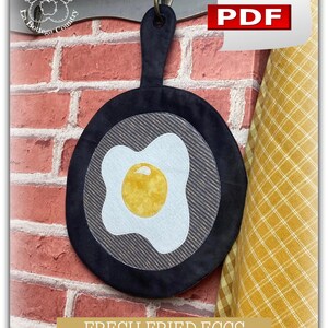 Peut inclure: Une poêle à frire en tissu matelassé noir et blanc avec un œuf blanc et jaune à l'intérieur. La poêle est accrochée à un mur de briques avec un tissu à carreaux jaune et blanc en arrière-plan. Le texte "FRESH FRIED EGGS" est sous la poêle.