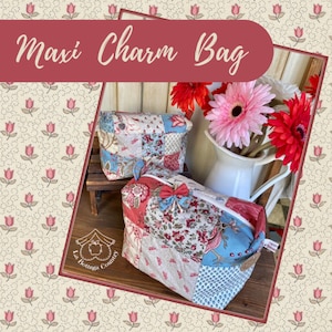 Può includere: Due pochette con cerniera in tessuto patchwork con un motivo floreale. Le pochette sono etichettate "Maxi Charm Bag" e sono appoggiate su una superficie in legno. Le pochette sono circondate da uno sfondo con un motivo floreale.