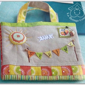 Könnte beinhalten: Eine beige Stofftasche mit einem gelben, grünen und orangefarbenen Streifenrand. Die Tasche zeigt eine Sonne mit einer Blume in der Mitte, eine Schmetterlingsapplikation und ein Banner mit der Aufschrift "It's Spring".