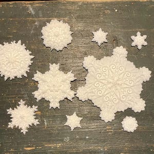 May include: A collection of eight white, three-dimensional snowflake ornaments of varying sizes. Each snowflake features intricate, embossed scrollwork and floral-inspired patterns across its surface, with distinct geometric shapes and pointed edges.