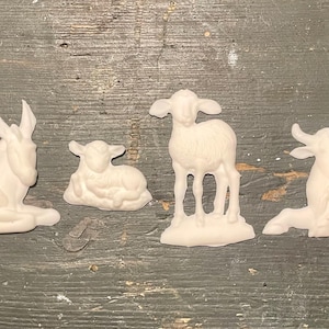 May include: A set of four unpainted white ceramic animal figurines including a donkey lying down, a small lamb lying down, a standing lamb, and a cow lying down. These matte white bisque pieces feature detailed textures representing fur and features.