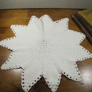 May include: White crocheted doily with a starburst design. The doily is made of delicate crochet stitches and has a circular center.