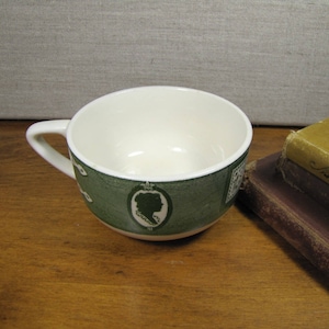 May include: A white teacup with a green and white floral design. The cup has a handle and is sitting on a wooden surface.