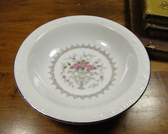 Può includere: Una ciotola in porcellana bianca con un motivo floreale con rose rosa e foglie verdi. La ciotola ha un bordo ondulato e un bordo blu.