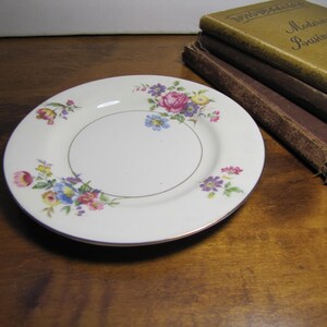 May include: A white ceramic plate with a floral design featuring pink, purple, and yellow flowers. The plate has a gold rim and is perfect for serving desserts or snacks.