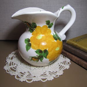 May include: A white ceramic pitcher with a yellow and green floral design. The pitcher has a wide mouth and a handle. It is sitting on a white crocheted doily.