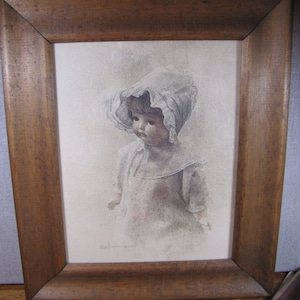 May include: A framed print of a young girl wearing a white bonnet and dress. The print is in a brown wooden frame.