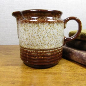 May include: A brown and white ceramic creamer with a textured glaze. The creamer has a rounded body and a handle.