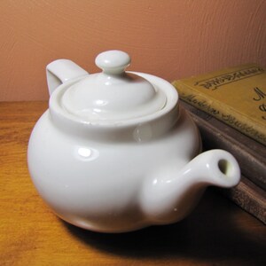 May include: A white ceramic teapot with a lid and a spout. The teapot is sitting on a wooden surface.