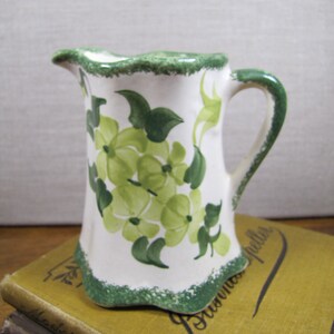 May include: A white ceramic pitcher with a green rim and floral design. The pitcher has a green floral pattern with white flowers and green leaves. The pitcher has a handle and a scalloped rim.