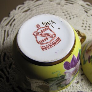May include: A yellow teacup with a floral design and a gold rim. The bottom of the cup has a red and white logo that reads "Bone Clarence China Made in England".