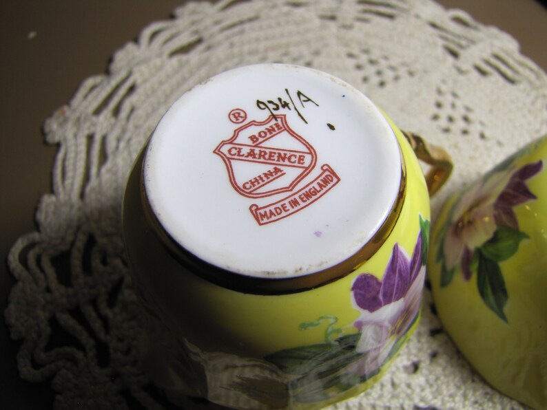 May include: A yellow teacup with a floral design and a gold rim. The bottom of the cup has a red and white logo that reads "Bone Clarence China Made in England".
