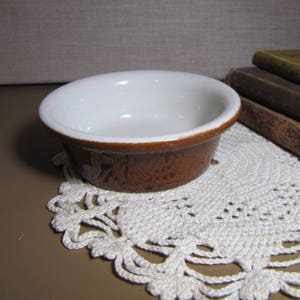 May include: A small, white ceramic bowl with a brown rim and a brown speckled exterior. The bowl is sitting on a white crocheted doily.