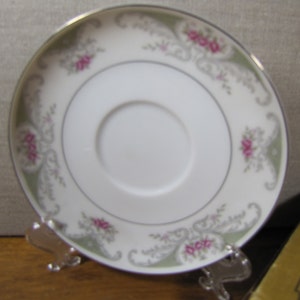 May include: A white saucer with a green and gray floral pattern and pink flowers. The saucer has a gold rim.