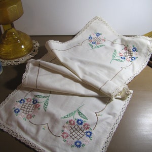 May include: White embroidered table runner with a crocheted edge. The runner features a floral design with pink, blue, and green flowers.
