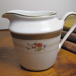 May include: A white porcelain creamer with a gold trim and floral design. The creamer has a small handle and a rounded body.