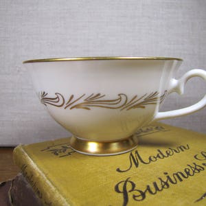 May include: White teacup with a gold rim and a gold floral design. The teacup is sitting on a stack of books with the text "Modern Business Speller" visible.