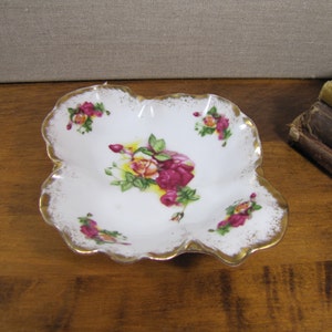 May include: A small, white porcelain dish with a scalloped edge and a gold trim. The dish is decorated with a floral pattern of pink and yellow roses.