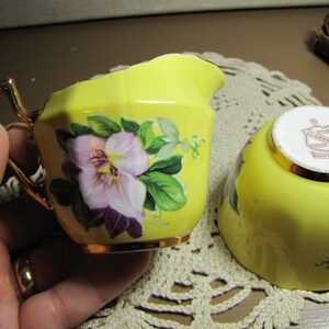 May include: A yellow creamer and sugar bowl set with floral designs. The creamer has a gold handle and a gold rim. The sugar bowl has a gold rim and a red and gold label that reads "Royal Clarence China Made in England".