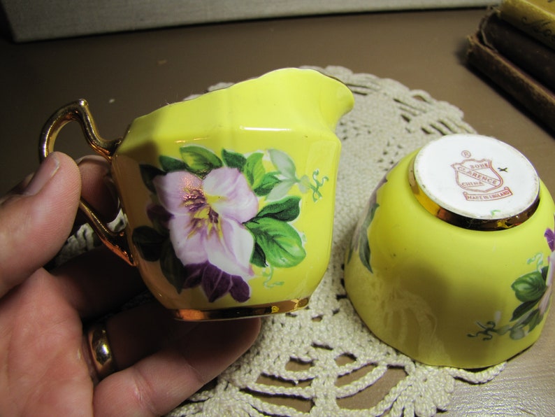 May include: A yellow creamer and sugar bowl set with floral designs. The creamer has a gold handle and a gold rim. The sugar bowl has a gold rim and a red and gold label that reads "Royal Clarence China Made in England".