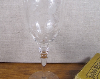 Può includere: Bicchiere da vino in vetro trasparente con disegno floreale inciso. Il bicchiere ha una base rotonda, un lungo stelo e una coppa ampia. Il disegno inciso presenta foglie e fiori. Il bicchiere è su una superficie di legno.